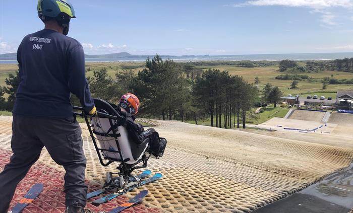A ski instructor holds a sit ski on top of an artificial slope with views of the coastline